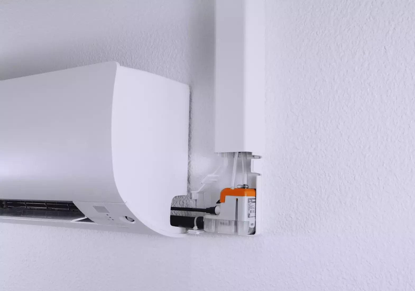 A close-up view of a white air conditioning unit installed in a corner, showing its upper section and attached piping along the wall with a mini condensate pump inside.